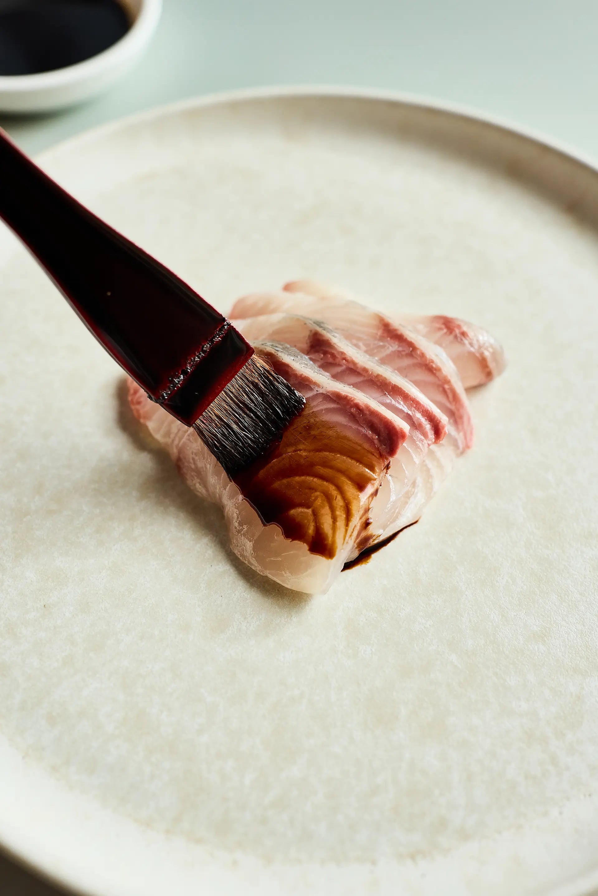 Shoyu being brushed on light colored fish on a stone plate with a dark colored brush