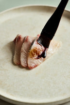 Sliced fish being brushed with dark shoyu on a light-colored plate.