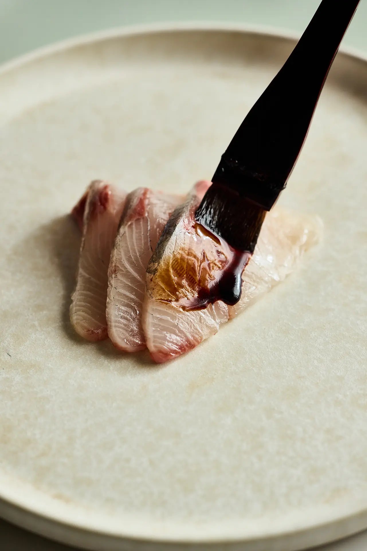 Sliced fish being brushed with dark shoyu on a light-colored plate.