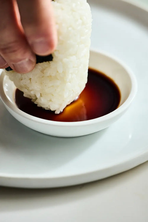 Musubi being dipped into marudaizu shoyu in a white ceramic dish