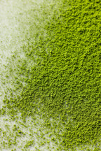 A close-up of Kobataen Koto matcha powder spread across a white surface, showing its vibrant green color and fine, soft texture with denser powder on the right and lighter dusting on the left.