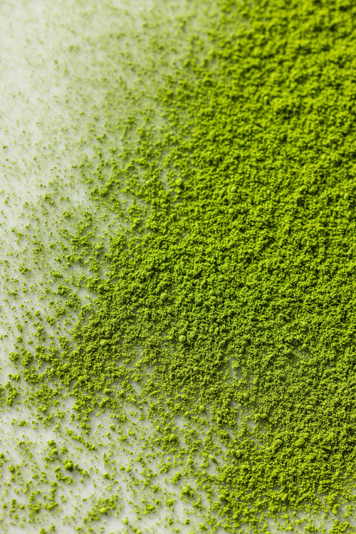 A close-up of Kobataen Koto matcha powder spread across a white surface, showing its vibrant green color and fine, soft texture with denser powder on the right and lighter dusting on the left.