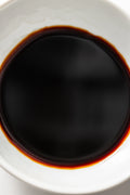 A white ceramic bowl filled with a dark, glossy liquid identified as Matsuai 10‑Year‑Aged Soy Sauce, showing a deep brown‑black color with a warm reddish tint around the edges.