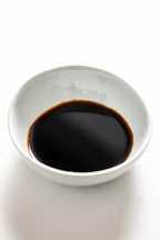 A white ceramic bowl filled with a dark, glossy liquid identified as Matsuai 10‑Year‑Aged Soy Sauce, showing a deep brown‑black color with a warm reddish tint around the edges.