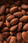 A close-up view of roasted almonds with dark brown, textured surfaces and visible ridges, lightly coated with coarse salt crystals.