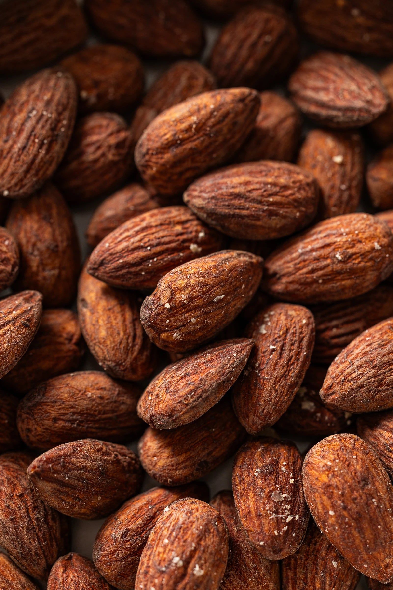 A close-up view of roasted almonds with dark brown, textured surfaces and visible ridges, lightly coated with coarse salt crystals.