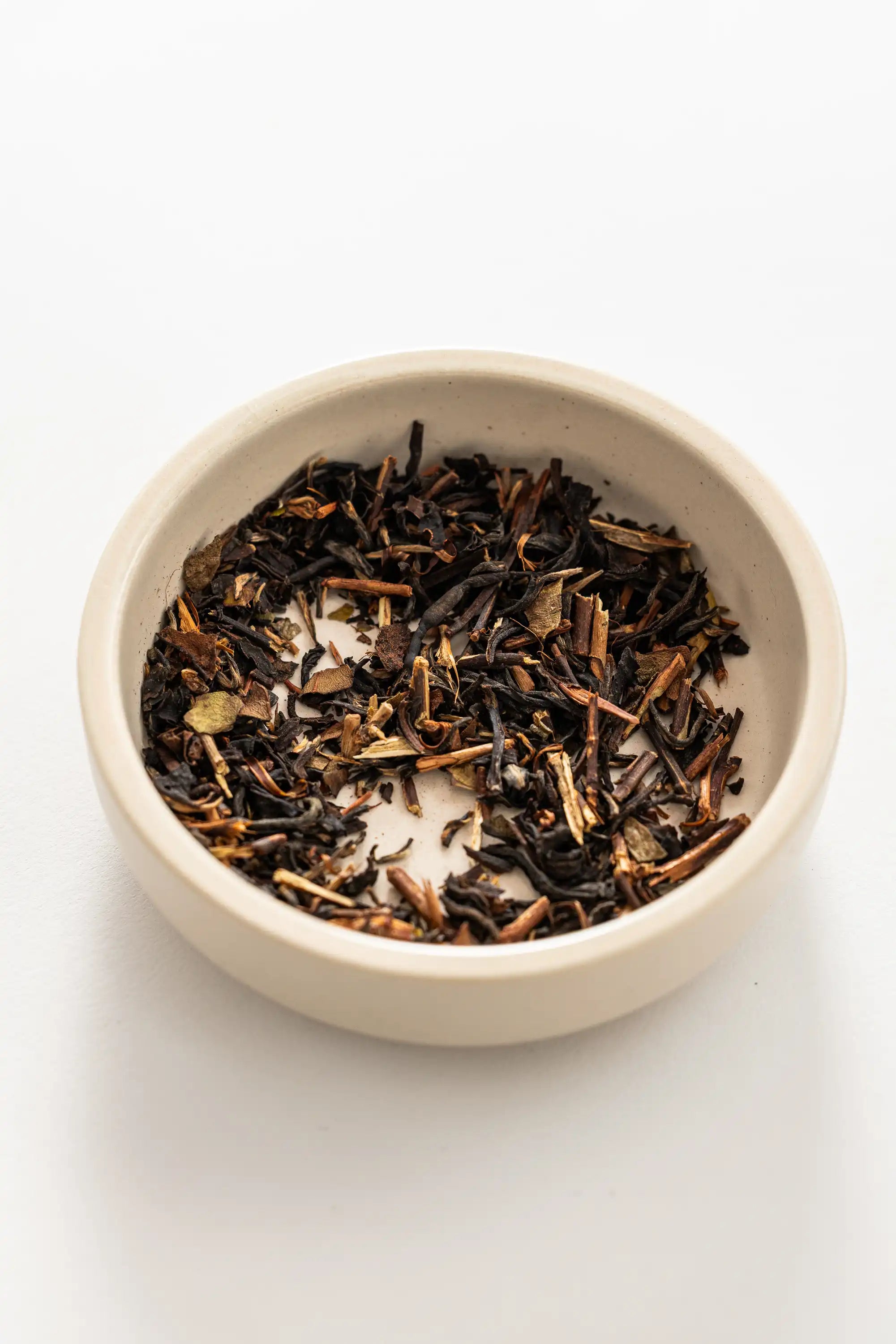 A small off‑white ceramic bowl filled with loose Oishi Farm black tea, showing dark brown to black twisted leaves mixed with lighter stems, creating a coarse, varied texture against a white background.