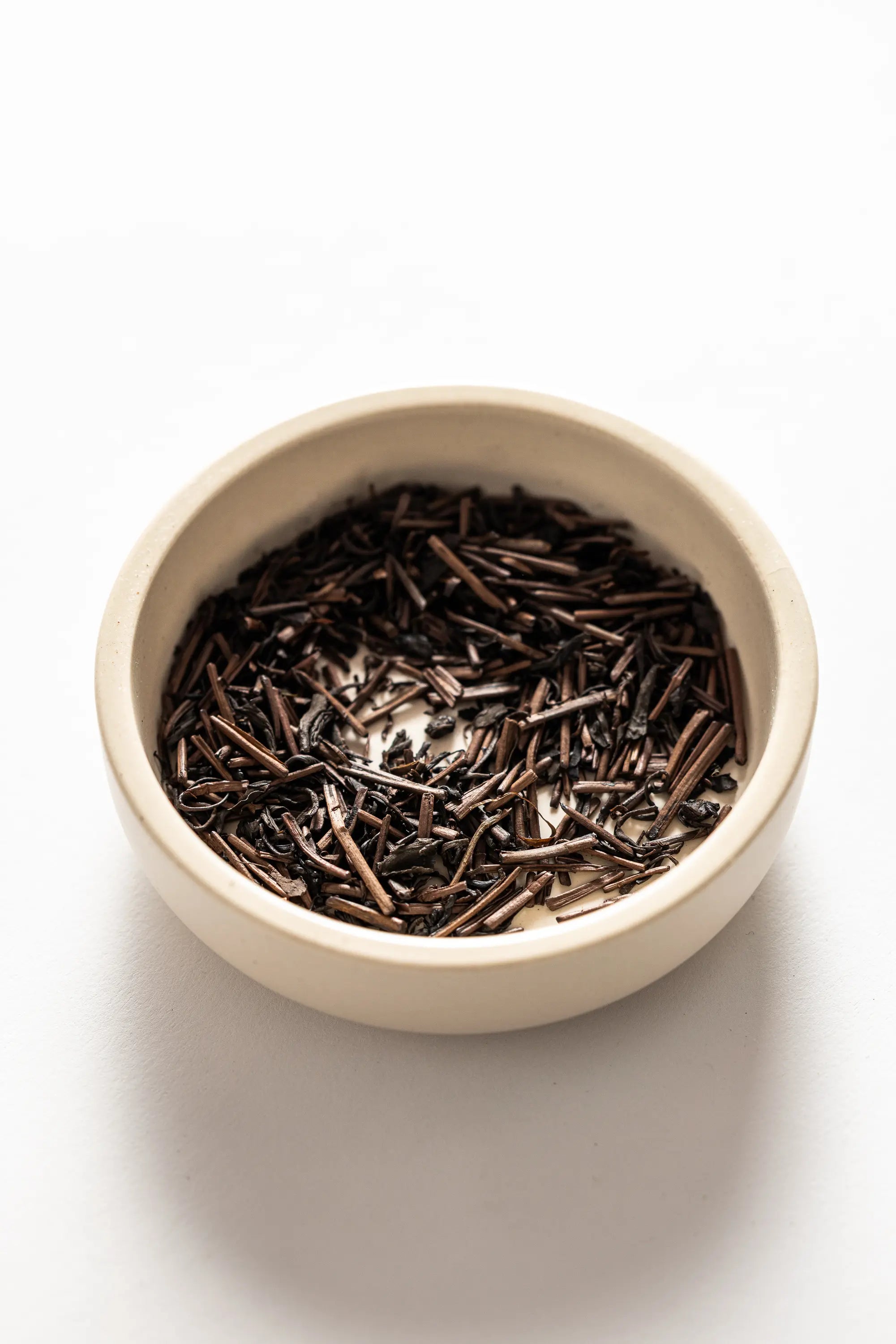 A close-up of Oyama Seicha hojicha, showing roasted brown tea leaves and stems with a mix of curled and twig-like pieces in a white dish on a white background.