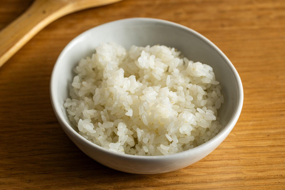A small white ceramic bowl filled with cooked white rice, placed on a warm wooden surface with a wooden spoon partially visible in the background.