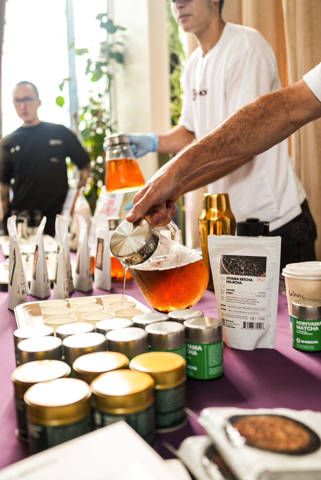 A table at an event displays various Japanese food products including jars, bags, and a cup, with a person pouring tea from a pitcher into a cup. Two people are visible in the background.