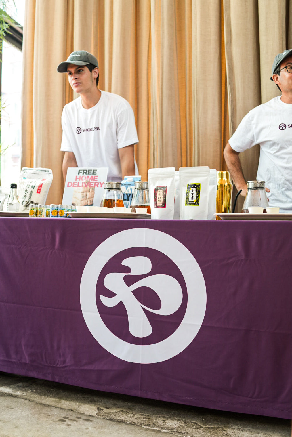 A pop up event scene featuring two men in white branded t-shirts behind a table draped with a purple cloth with a large white Shokuya logo. The table is stocked with various product containers, including bags and bottles, and is set against a backdrop of beige curtains.