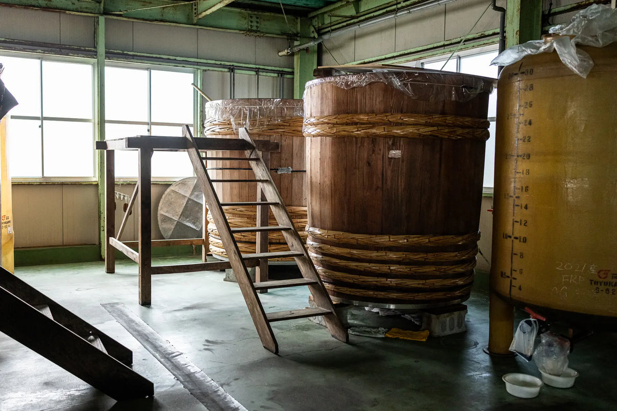 A large wooden kioke barrel with a wooden ladder leaning against it, set inside a warehouse or production room with windows, concrete floors, and other large vessels nearby.
