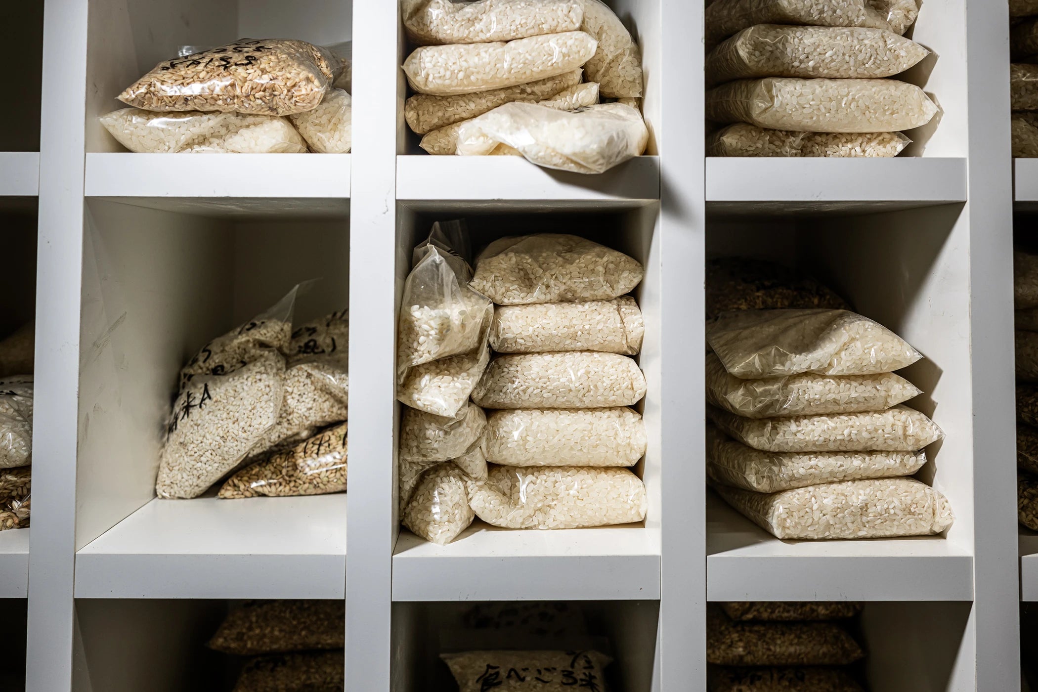Japanese rice grain variety samples displayed in bags on white shelving at Kanegae facility.