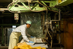 A factory worker wearing a white coat, apron, blue gloves, and a light green cap operates machinery while pressing sheets of material used in shoyu production. The worker positions the material on a metal machine surrounded by control panels, wiring, and industrial equipment.