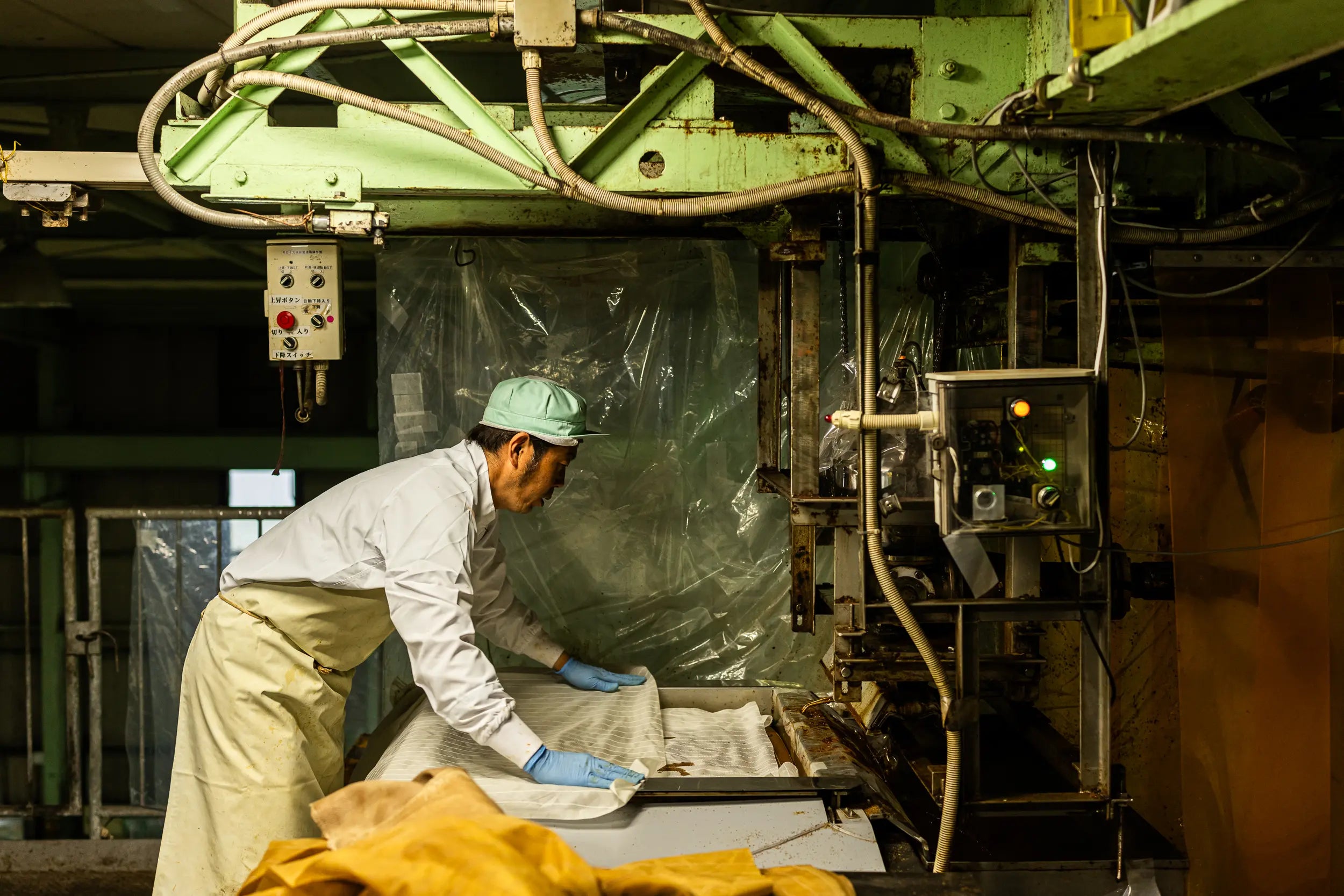A factory worker wearing a white coat, apron, blue gloves, and a light green cap operates machinery while pressing sheets of material used in shoyu production. The worker positions the material on a metal machine surrounded by control panels, wiring, and industrial equipment.