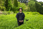 Person (Oyama-San) standing in a lush green tea field with trees in the background.
