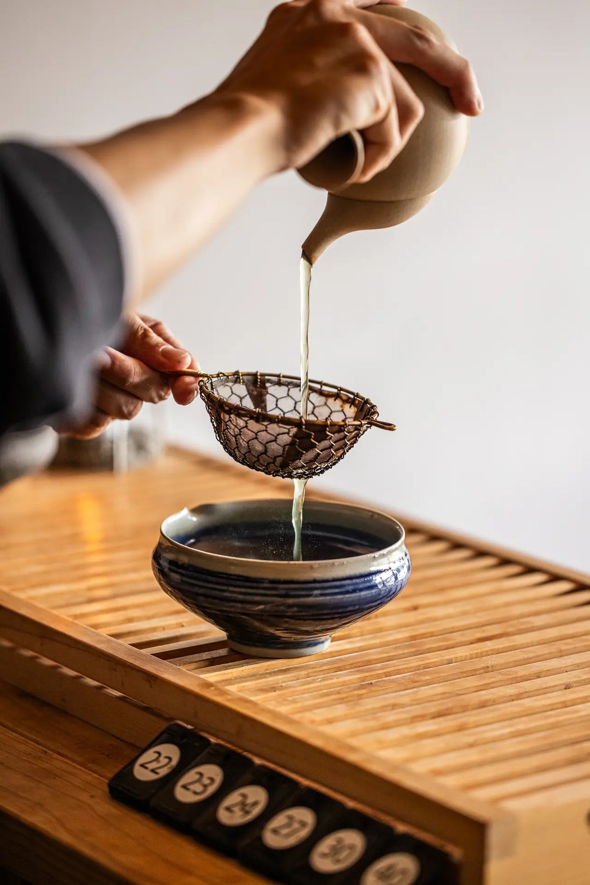 A close-up of tea being poured from a beige ceramic teapot through a metal strainer into a blue-and-white ceramic bowl on a wooden tea tray.