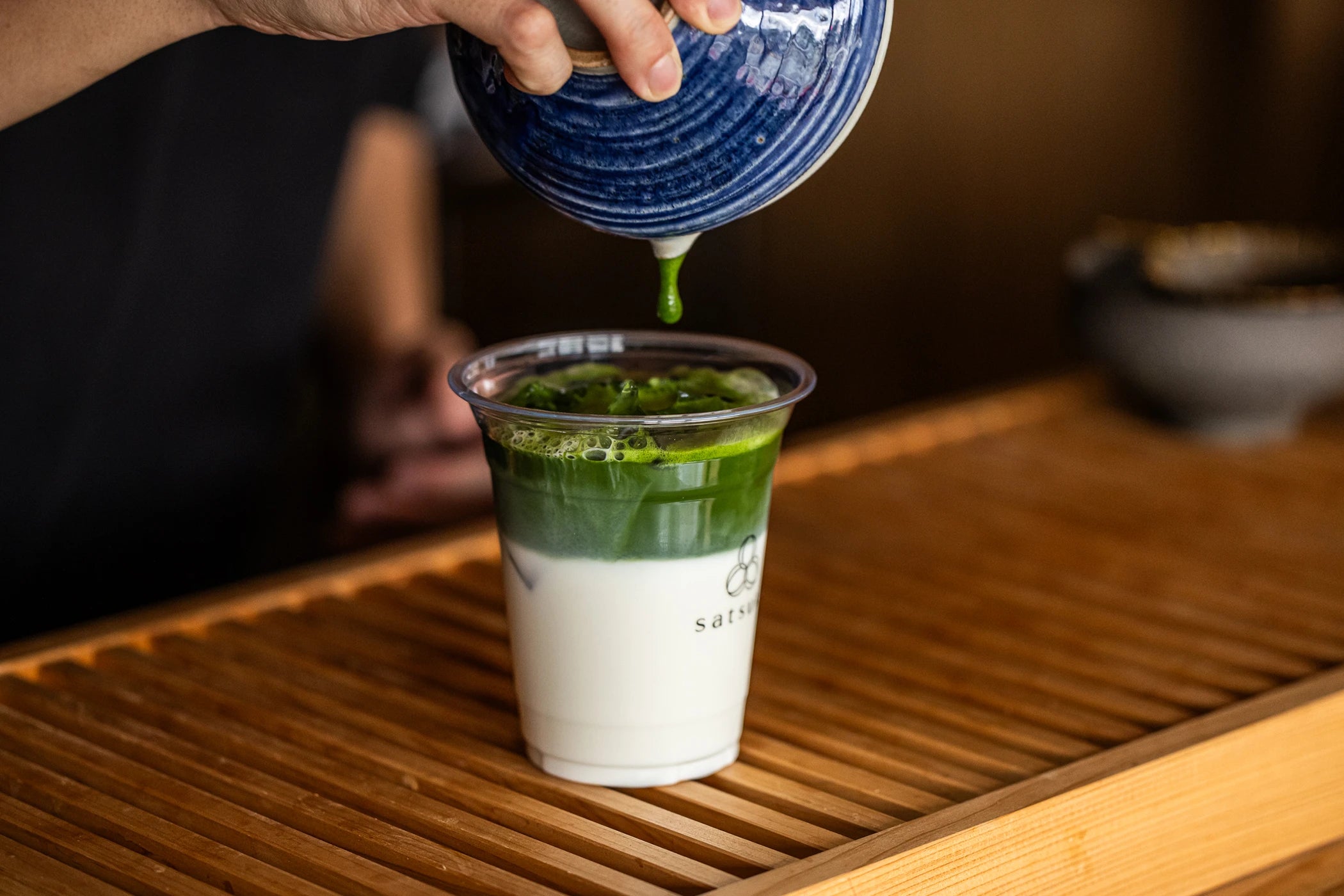 Person pouring whisked matcha tea into a cup of milk on a wooden tray.