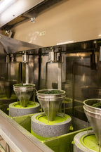 A metal matcha grinding machine with multiple stone mills, each topped by a funnel of green tea leaves. The stones rotate to grind the leaves into fine matcha powder, visible as green dust around the bases.