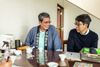 Two people sit at a table having a conversation. One wears traditional Japanese clothing with a dark sash featuring white characters, and the other wears glasses and a dark sweater. On the table are white ceramic tea cups, a teapot, a spray bottle, a clipboard with a pen, and a basket containing green packages and a black pouch.