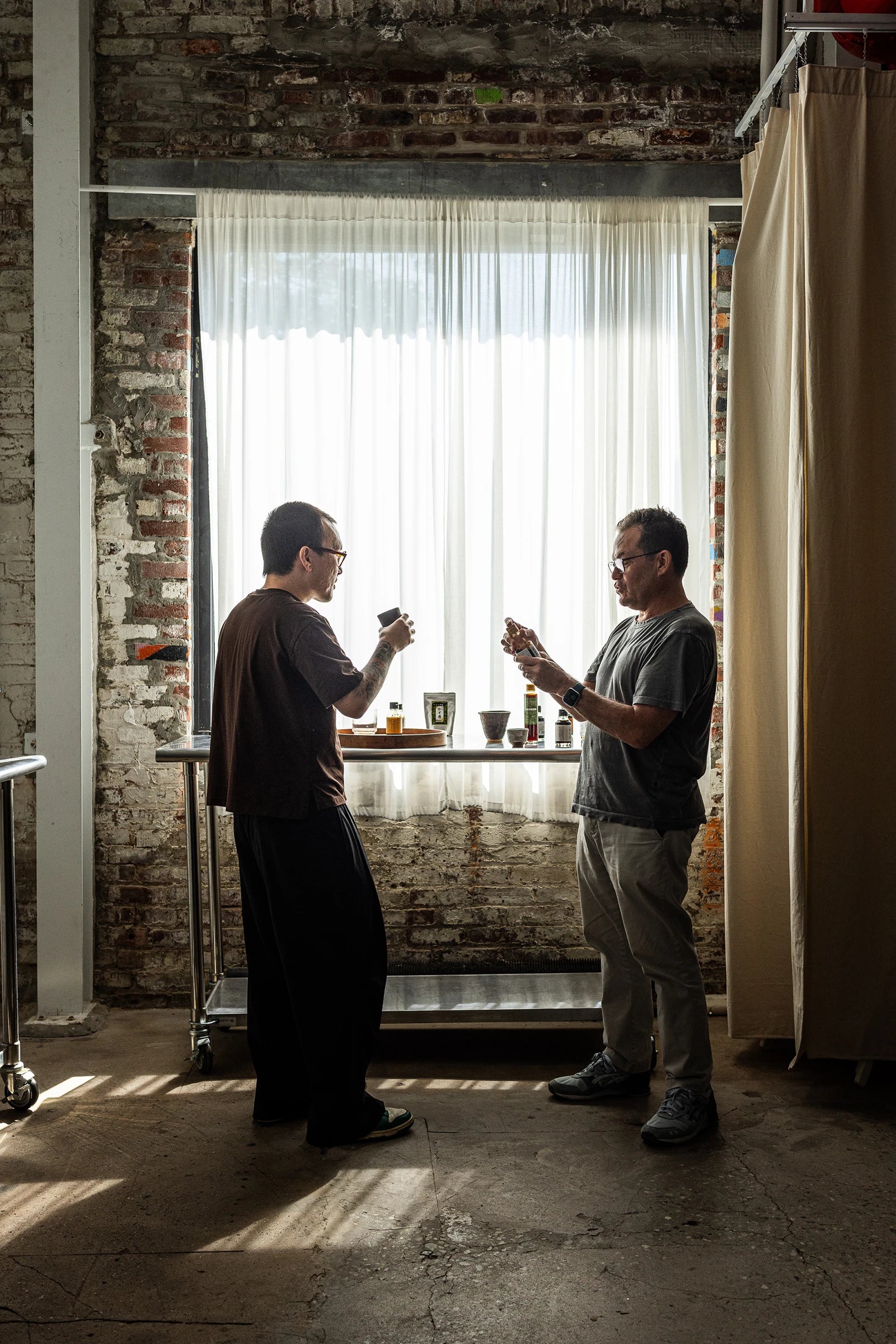 Two men standing in a room with brick walls and a large window, engaged in a conversation and tasting shoyu, soy sauce.
